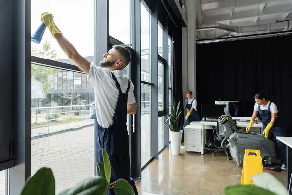 Bärtiger Mann wäscht Bürofenster in der Nähe von interrassischen Frauen, die auf verschwommenem Hintergrund arbeiten — Stockfoto