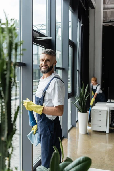 Cheerful bearded man with detergent and rag looking at camera near office windows — Stock Photo