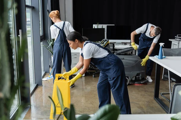 Équipe de nettoyeurs multiethniques et professionnels en uniforme travaillant dans un bureau moderne — Photo de stock