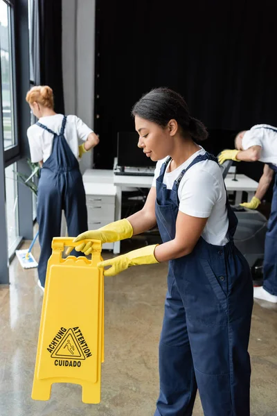 Young bi-racial woman holding warning board near colleagues cleaning office — Stock Photo