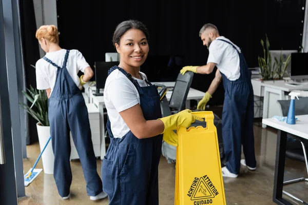 Heureuse femme bi-raciale avec panneau d'avertissement regardant la caméra près de collègues travaillant dans le bureau — Photo de stock