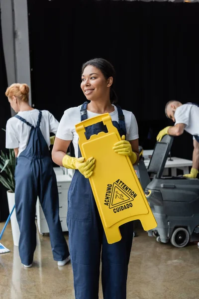 Feliz bi-racial mujer sosteniendo tablero de precaución cerca de compañeros de trabajo y máquina de fregar piso - foto de stock