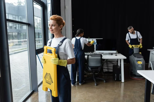 Sonriente mujer sosteniendo atención tablero cerca de ventanas mientras interracial colegas limpieza oficina - foto de stock