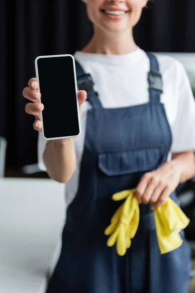 Частичный вид размытой женщины в комбинезоне с резиновыми перчатками и смартфоном с чистым экраном — стоковое фото