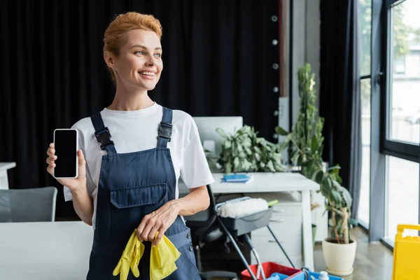 Donna allegra in tuta che tiene smartphone con schermo bianco mentre distoglie lo sguardo in ufficio — Foto stock