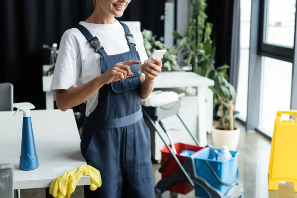 Vista ritagliata di donna sorridente utilizzando smartphone vicino a detergenti e guanti di gomma — Foto stock