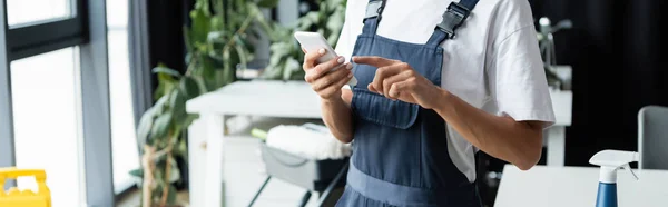 Vista recortada de limpiador profesional en overoles apuntando al teléfono móvil, bandera - foto de stock