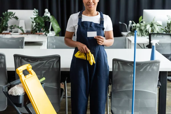 Partial view of smiling bi-racial woman with blank business card near cleaning supplies in office — Stock Photo