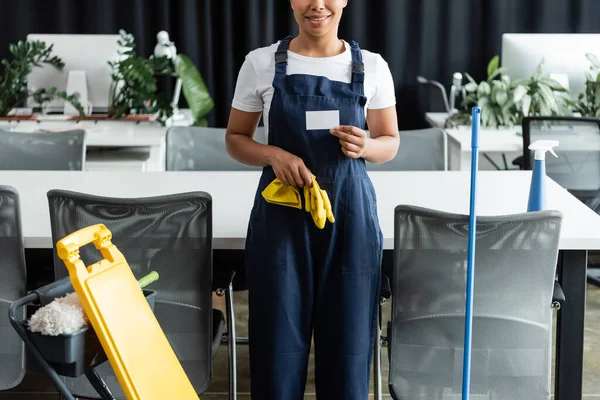 Vista parziale della donna sorridente bi-razziale con biglietto da visita vuoto vicino alle forniture per la pulizia in ufficio — Foto stock