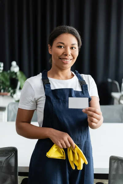 Симпатичная двурасовая женщина в форме держит резиновые перчатки и чистую визитку — стоковое фото