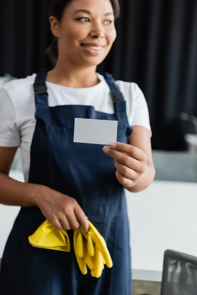 Улыбающаяся двурасовая женщина в резиновых перчатках держит пустую визитку и отворачивается — стоковое фото