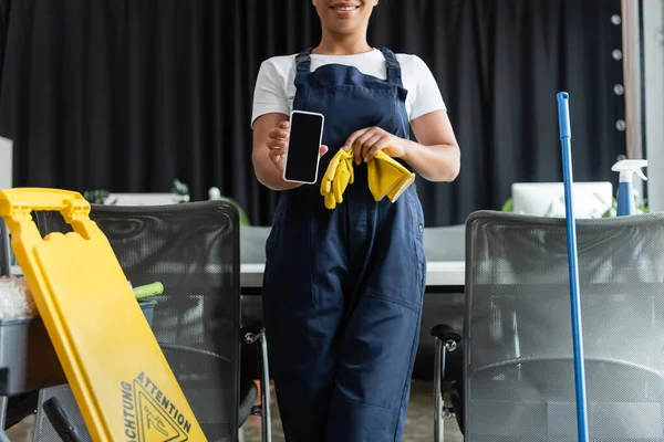 Partial view of smiling bi-racial woman in overalls showing smartphone with blank screen in office — Stock Photo