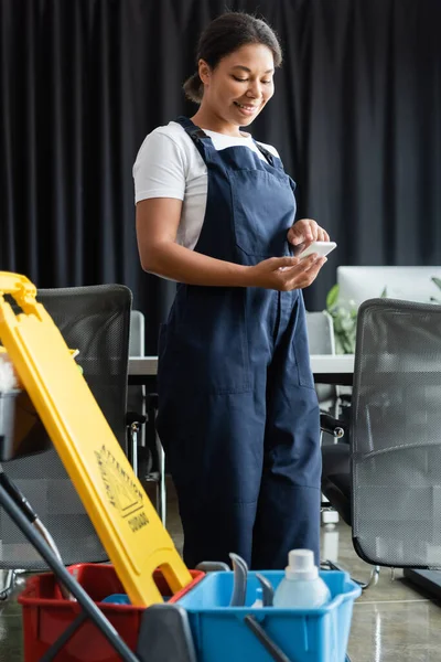 Cheerful bi-racial woman in workwear using smartphone near cart with cleaning supplies — Stock Photo