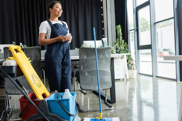 Glückliche zweirassige Frau, die mit Handy in der Nähe von Warntafel und Einkaufswagen mit Putzmitteln steht — Stockfoto