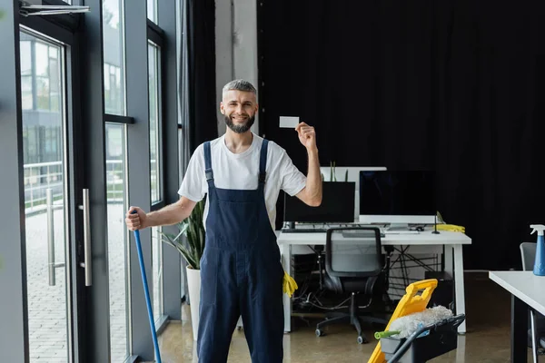 Glücklicher Mann blickt in die Kamera und zeigt leere Visitenkarte neben Einkaufswagen mit Putzmitteln — Stockfoto