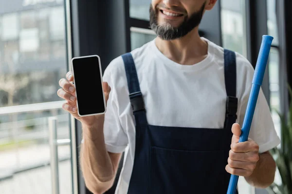 Partial view of happy bearded cleaner in overalls holding mobile phone with blank screen — Stock Photo