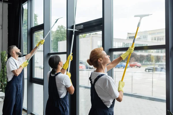 Limpadores profissionais multiétnicos em lavagem uniforme grandes janelas no escritório — Fotografia de Stock