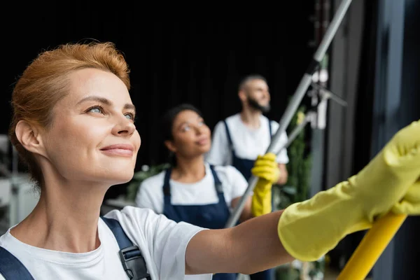 Foco seletivo da mulher feliz lavando janelas perto da equipe de limpeza multiétnica borrada — Fotografia de Stock
