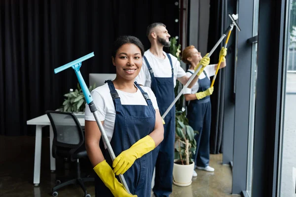 Joyeuse femme bi-raciale tenant essuie-glace près de collègues travaillant dans le bureau — Photo de stock