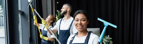 Junge und glückliche Putzfrau, die in die Kamera lächelt, während Kollegen Fenster waschen, Banner — Stockfoto