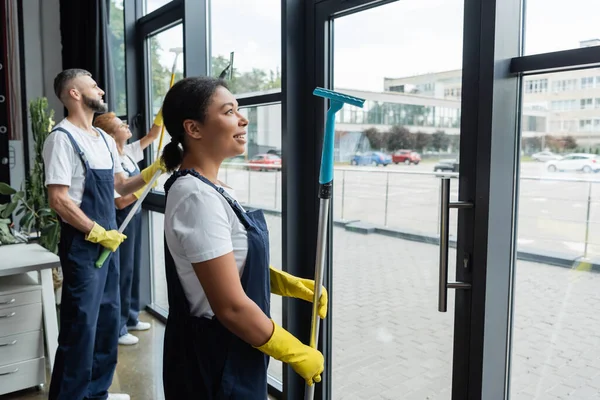 Jeune femme bi-raciale en vêtements de travail souriant près des collègues laver les fenêtres du bureau — Photo de stock