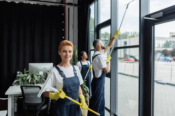 Femme souriante en gants de caoutchouc regardant la caméra près de collègues multiethniques nettoyer de grandes fenêtres de bureau — Photo de stock