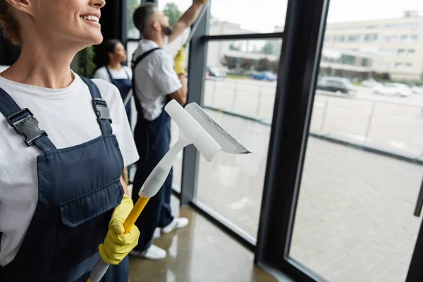 Smiling woman in overalls holding window squeegee near blurred multiethnic colleagues — Stock Photo