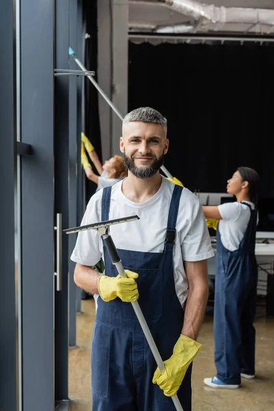 Glücklich bärtige Putzfrau mit Scheibenwischer lächelt in die Kamera neben interrassischen Frauen, die im Büro arbeiten — Stockfoto