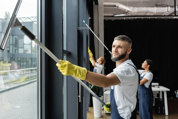 Bearded man in rubber gloves washing windows near interracial team in office — Stock Photo