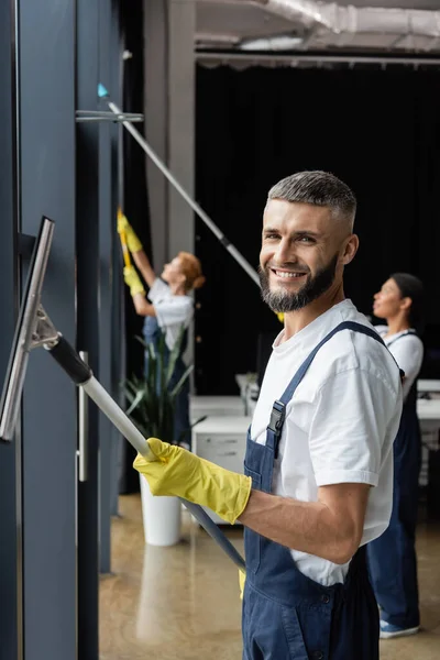 Bearded man looking at camera while washing windows near multiethnic colleagues on blurred background — Stock Photo