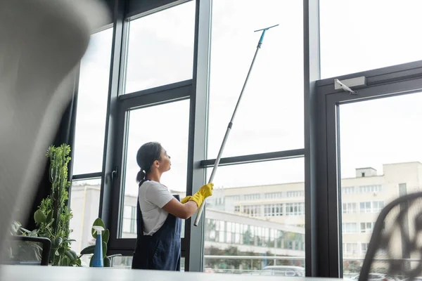 Femme bi-raciale en tenue de travail lavant de grandes fenêtres de bureau au premier plan flou — Photo de stock