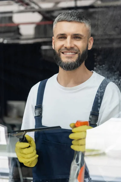 Limpador barbudo com detergente e limpador de janela sorrindo para a câmera perto de vidro molhado — Fotografia de Stock