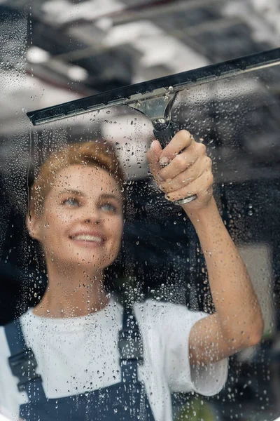 Professional cleaner with window squeegee smiling near wet glass in office — Stock Photo