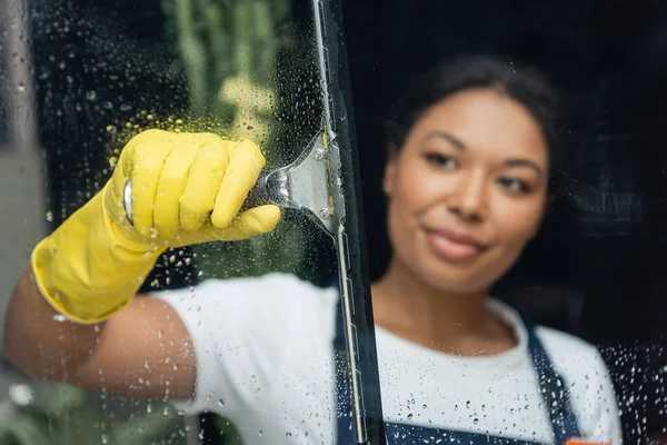 Femme bi-raciale floue en gant de caoutchouc nettoyage verre avec essuie-glace — Photo de stock