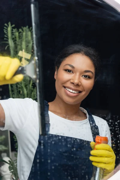 Bi-racial feliz mujer en guantes de goma mirando a la cámara mientras se lava ventana en la oficina - foto de stock