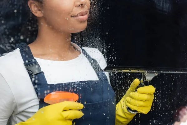 Vista recortada de la mujer bi-racial con detergente y escobilla de ventana cerca de vidrio húmedo en la oficina - foto de stock