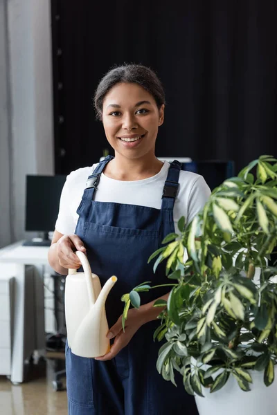 Feliz bi-racial mujer con riego puede mirar a la cámara cerca de planta verde - foto de stock