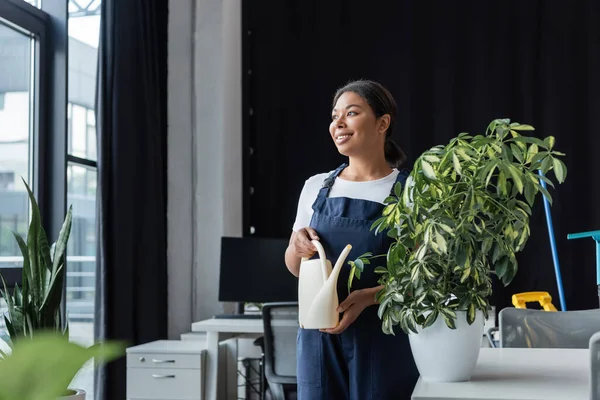 Heureuse femme bi-raciale en tenue de travail debout avec arrosage peut près de la plante et regardant loin — Photo de stock