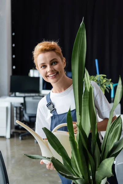 Fröhliche professionelle Reinigungskraft mit Gießkanne blickt in die Kamera in der Nähe der grünen Pflanze — Stockfoto