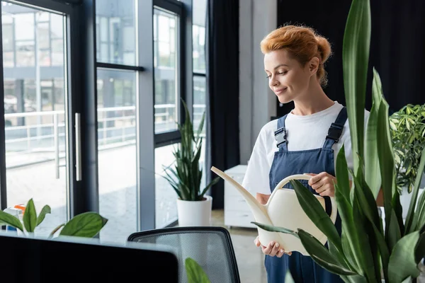 Lächelnde Frau in Overalls mit Gießkanne in der Nähe grüner Pflanze im Büro — Stockfoto