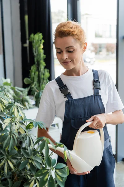 Femme souriante en salopette arrosage plante verte au bureau — Photo de stock