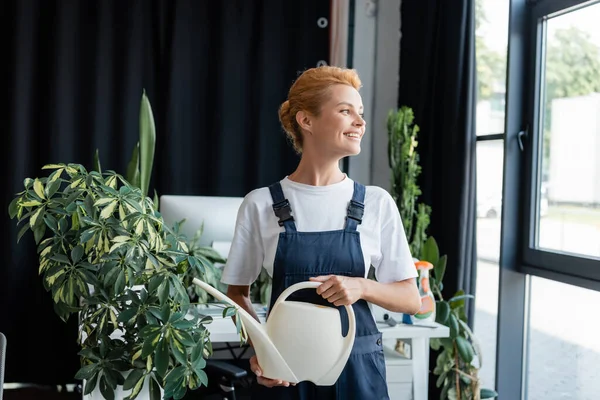 Mujer feliz con regadera puede mirar hacia otro lado mientras está de pie cerca de la planta en la oficina — Stock Photo