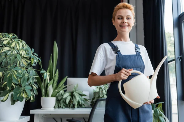 La donna allegra con annaffiamento può guardare la macchina fotografica vicino a impianti in vaso in ufficio — Foto stock