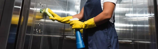 Vista recortada de limpiador bi-racial en ropa de trabajo y guantes de goma limpieza oficina ascensor, pancarta - foto de stock