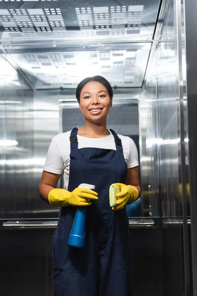 Jovem bi-racial mulher em macacão segurando spray garrafa e esponja no elevador — Fotografia de Stock