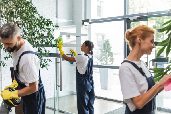 Equipo multiétnico de limpiadores profesionales en overoles lavando vestíbulo de la oficina - foto de stock