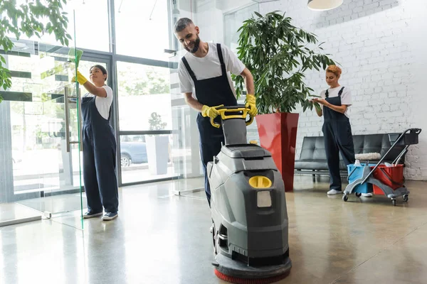 Man in workwear with floor scrubber machine near interracial women cleaning office lobby — Stock Photo