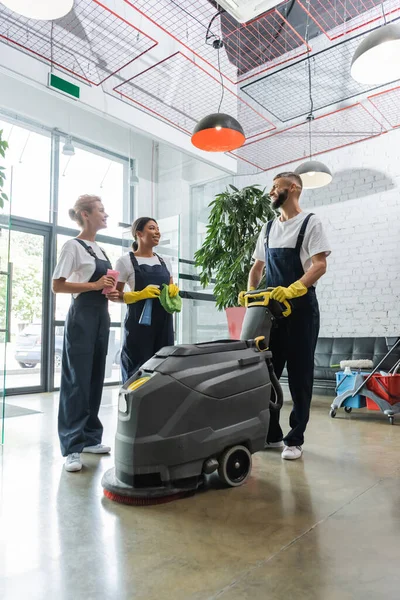 Happy multiethnic professional cleaners talking near floor scrubber machine in office lobby — Stock Photo