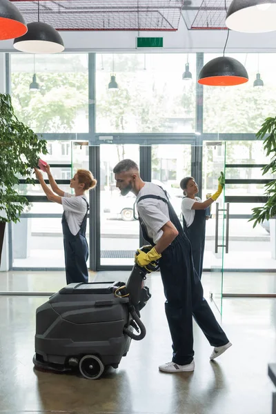 Side view of man with floor scrubber machine near multicultural women cleaning office hall — Stock Photo