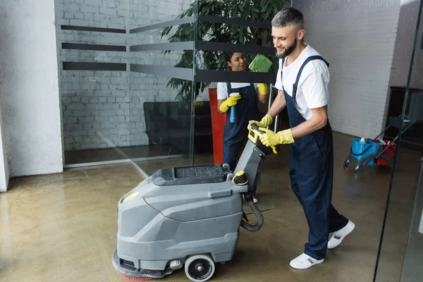 Bearded man with floor scrubber machine near bi-racial woman washing glass door in office — Stock Photo